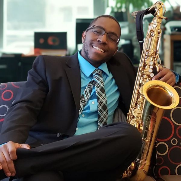 Ben Green An image of Ban Green with his saxophone sitting on a couch with a huge smile wearing a dark suit and a blue shirt with a patterned tie.